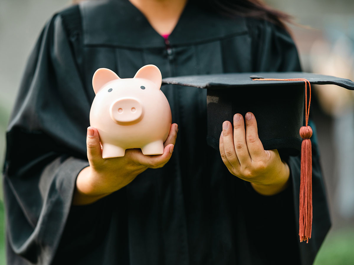A graduate in a black robe holds a piggy bank in one hand and her graduation cap in the other.
