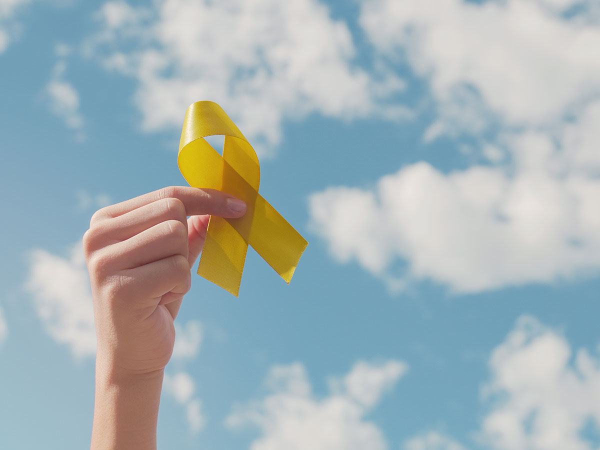 A hand holding up a yellow ribbon against a blue sky with scattered clouds. 