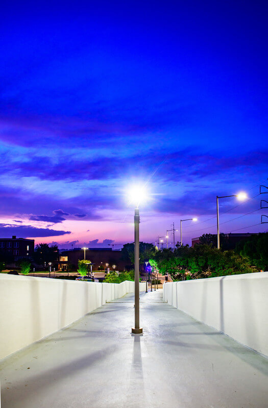 A UAB pedestrian bridge at night, an illuminated lamp in the middle, under a violet and pink sky. 
