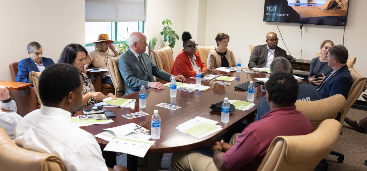 Watts sitting at a conference table with many others.
