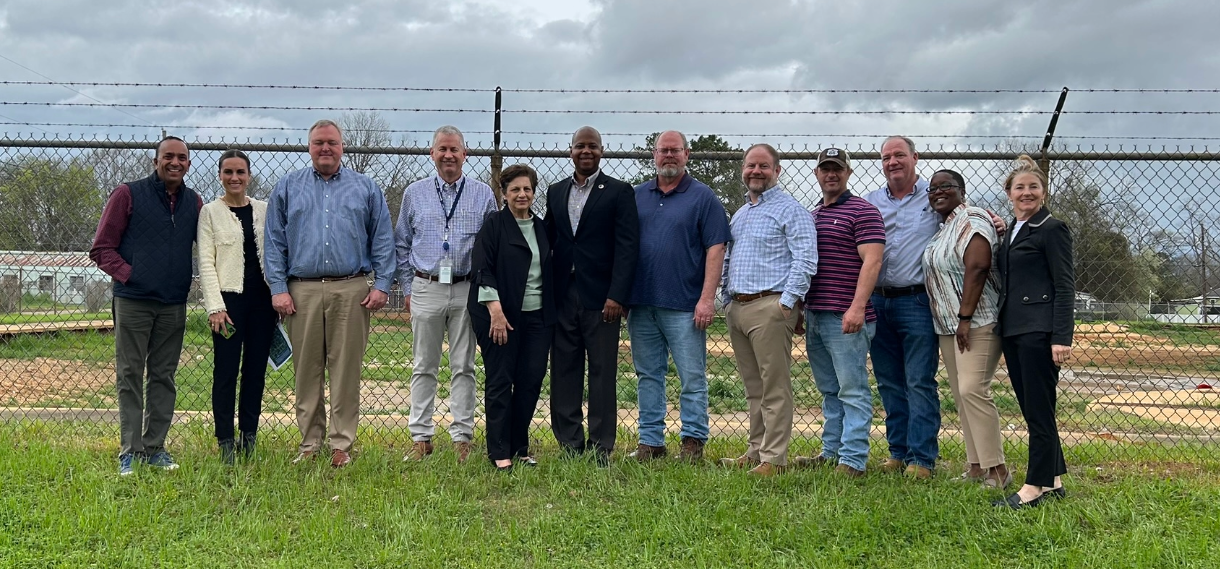 A group of people standing on the grass in front of a chain link fence.