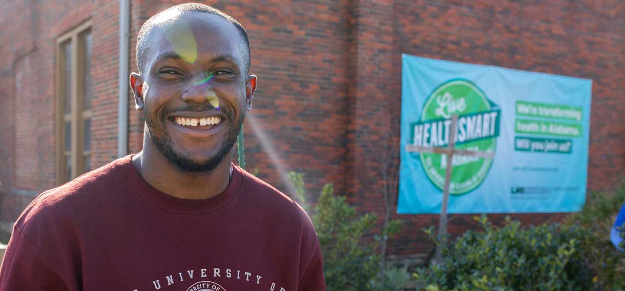A young black man smiles while standing next to Live HealthSmart banner on the side of a brick building.