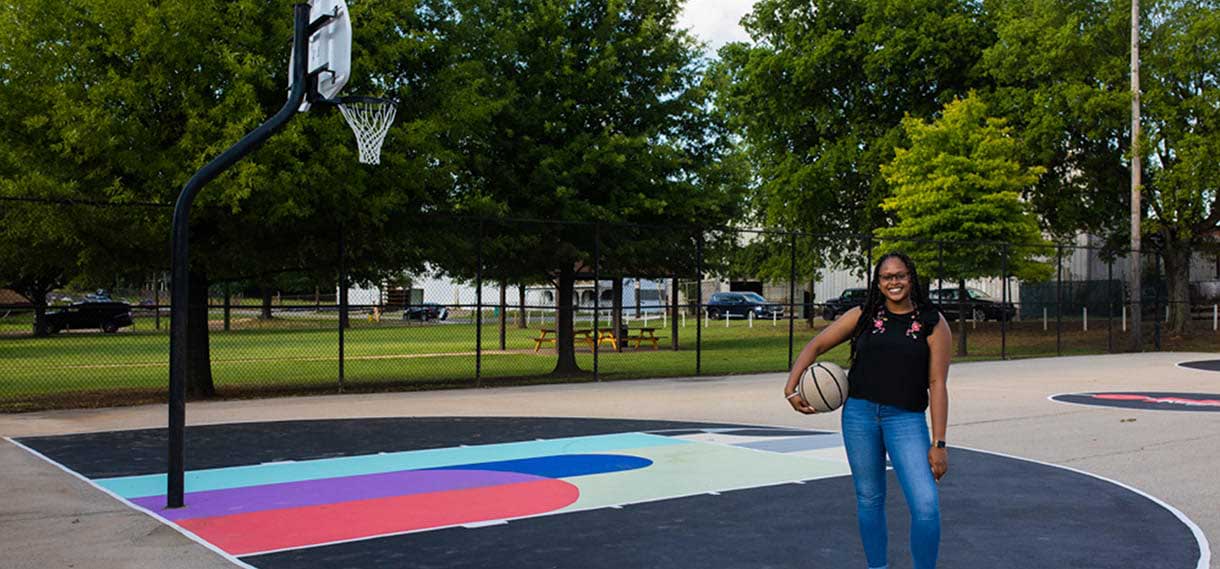Black woman holding a basketball on the park's colorful outdoor basketball court.