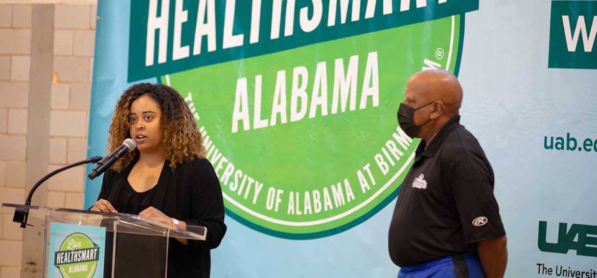 A Black woman speaks at a podium while a Black man looks on.