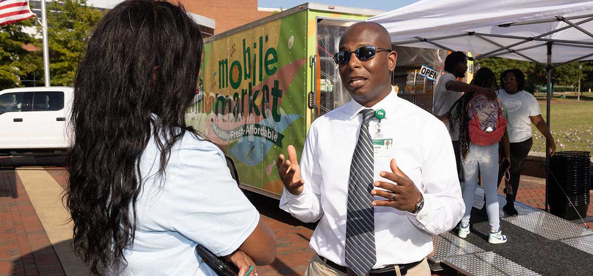 A Black man in a white dress shirt and tie talks to a Black woman outside the mobile market.