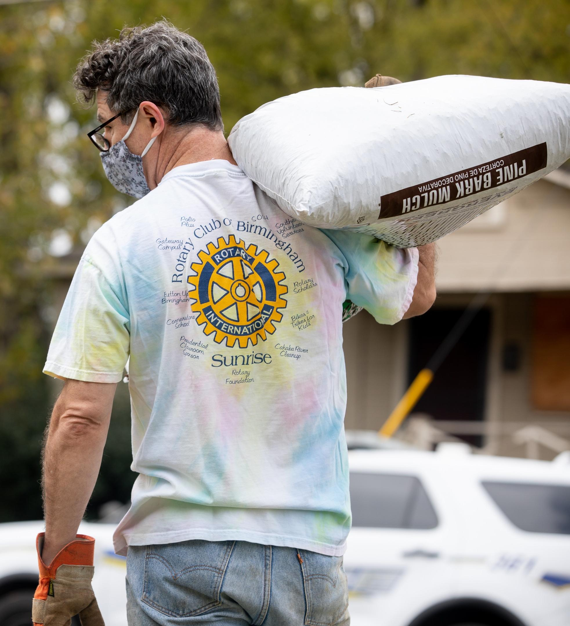 A volunteer in a tie-dye rotary shirt and work gloves carries a large bag of pine bark mulch. 