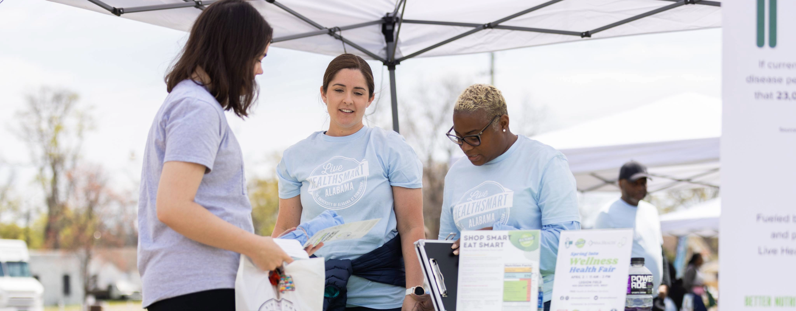 Young woman in a lavender tshirt talks to two women at an outdoor Live HealthSmart info table. 