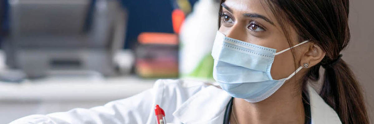 A female doctor bend's down to eye level as she talks with a cancer patient who is receiving her treatment intravenously.  She is wearing a white lab coat and has a clipboard in hand as she takes notes.  The patient is dressed casually and she sits back in the chair waiting for her treatment to finish, and both are wearing medical masks to protect them from COVID.