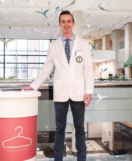 Nik Hakes poses with a collection bin for Compassion Closet in the lobby of UAB hospital. The collection bin is marked with a hanger logo on a pink background. 