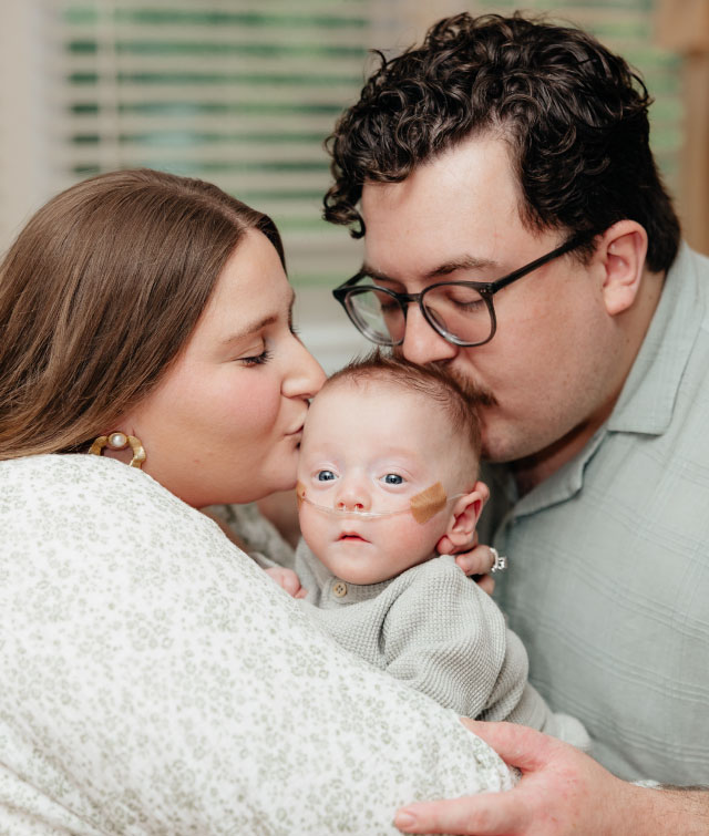 A mother and father kiss their infant son while holding the baby between them. The baby, wearing a light-colored sweater and with a nasal cannula secured with medical tape, looks toward the camera.