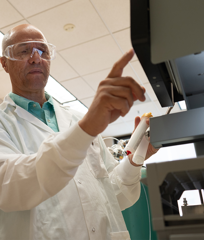 Research manager Omar Moukha-Chafiq wearing a lab coat and safety glasses operates laboratory equipment using a touchscreen while holding a sample container.