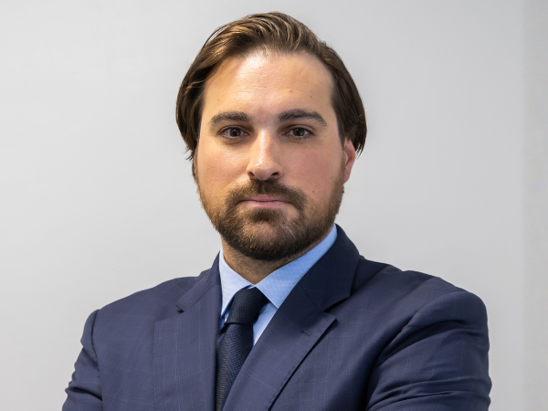 Professional headshot of a person wearing a navy blue suit, light blue dress shirt, and dark tie against a light gray background.