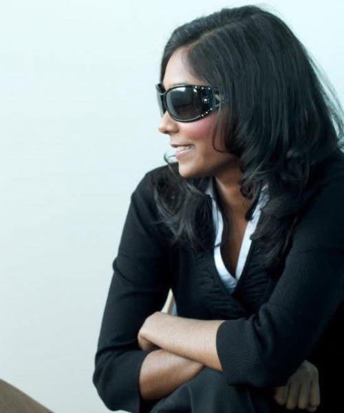 Person wearing dark sunglasses and a black jacket, seated with folded arms against a light background