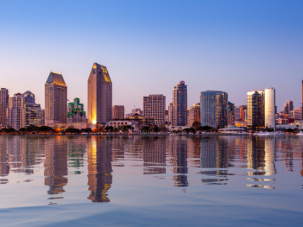 Skyline of downtown San Diego at dusk, with high-rise buildings illuminated against a blue and pink evening sky. The city lights reflect across calm water in the foreground, creating mirrored vertical streaks on the surface.