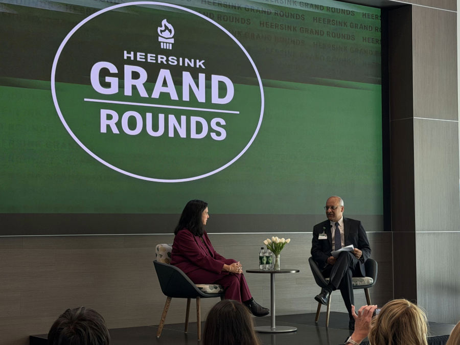 Sharmila Makhija, M.D., MBA,  and Anupam Agarwal, M.D., sit on a stage. A large green graphic behind them displays "Heersink Grand Rounds" in white text with a white torch and a white circle.