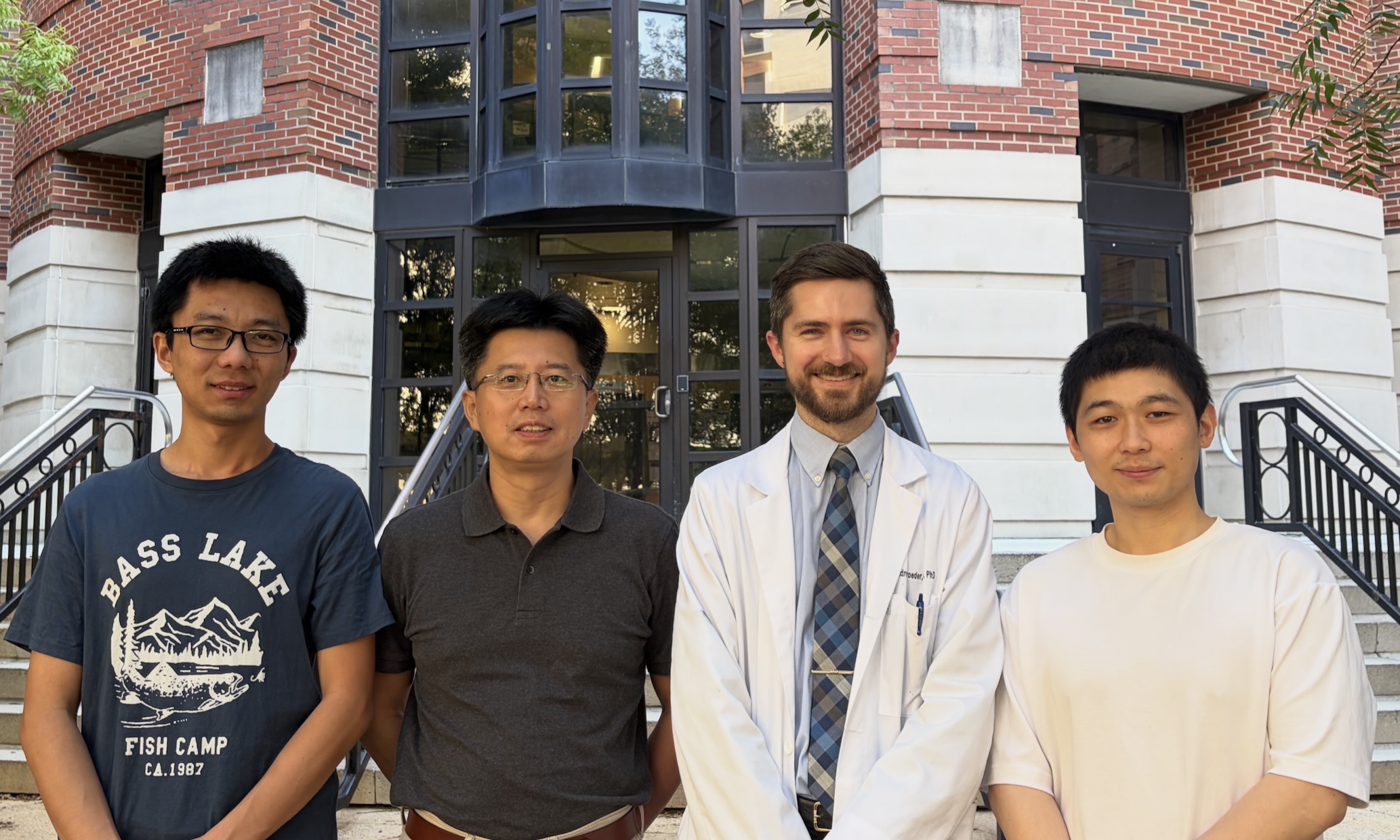 Four individuals stand side by side outdoors in front of a red brick academic building with large windows and stair railings. One person wears a white lab coat and tie, while the others wear casual shirts. The group is centered in front of the building entrance during daylight.