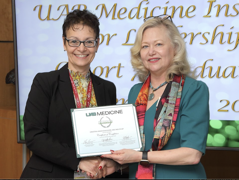 Dr. Magi-Galluzzi (left) receives a framed certification from Dr. Larson (right) during a recognition event. Both stand smiling and holding the certificate together in front of a presentation screen displaying event branding for a UAB Medicine institute program.