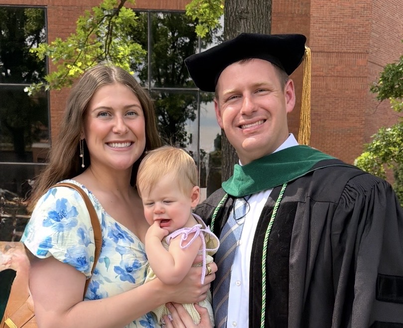 Outdoor graduation portrait of three people standing together in front of a brick building and trees. One adult wears academic regalia including a black doctoral-style gown, dark academic cap with gold tassel, and a green hood over a dress shirt and tie. Another adult stands beside them wearing a light blue and white floral dress and shoulder bag. They are holding a young child dressed in a light-colored outfit. All three are close together, creating a warm family celebration moment in natural daylight.