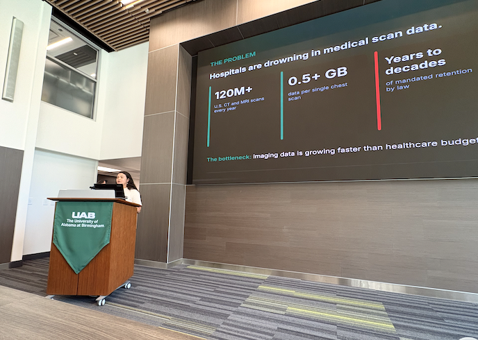 A presenter speaks at a podium in a UAB conference space while a large presentation slide behind them highlights challenges in medical imaging data growth. The slide includes statistics on annual U.S. CT and MRI scans, data generated per chest scan, and long-term data retention requirements, emphasizing pressures on healthcare infrastructure. The room features contemporary wood paneling, high ceilings, and a large digital display.
