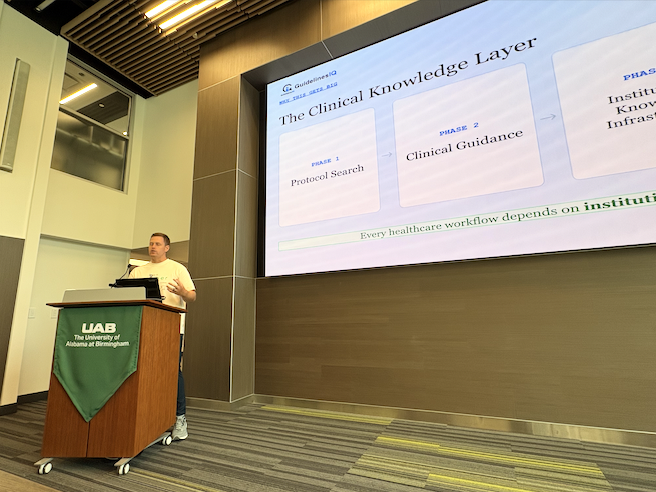 A presenter stands at a podium in a modern lecture hall at the University of Alabama at Birmingham while delivering a presentation. A large projected slide behind them displays a framework titled “The Clinical Knowledge Layer,” with sections labeled Protocol Search and Clinical Guidance. The podium features a green UAB banner, and the room includes tall ceilings, wood accents, and tiered lighting.