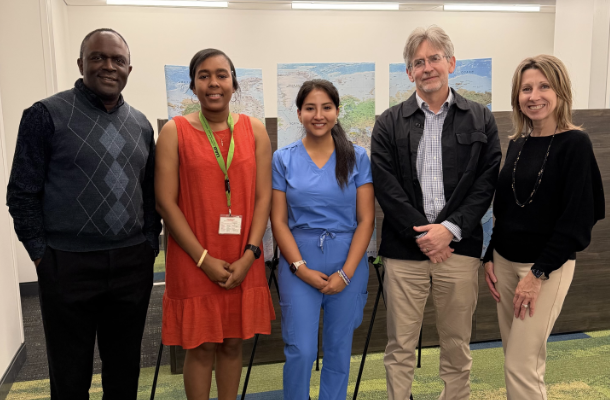 Group photo of five people standing together indoors in a academic setting. The group includes individuals dressed in business casual attire and one person in blue medical scrubs. Behind them are display boards featuring landscape imagery. The group is standing close together and smiling toward the camera in a bright meeting or collaboration space.