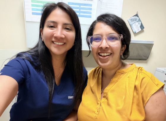 Casual indoor photo of two people standing together in a clinical or exam room setting. One person is wearing navy blue medical scrubs, and the other is wearing a mustard-yellow blouse and glasses. Both are smiling toward the camera. Medical equipment, posted materials, and cabinets are visible in the background, creating a warm and informal healthcare environment.