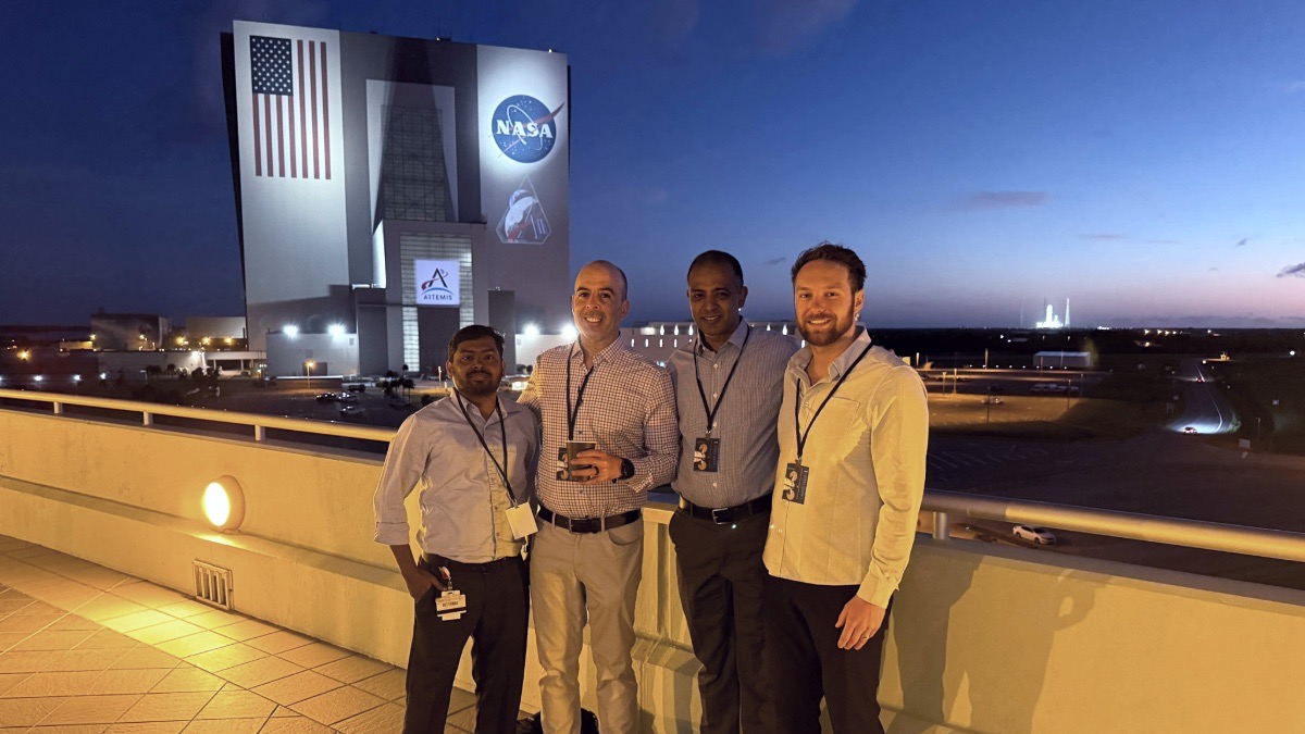 Four people with event badges stand on a rooftop at dusk, with NASA’s Vehicle Assembly Building and U.S. flag visible in the background.