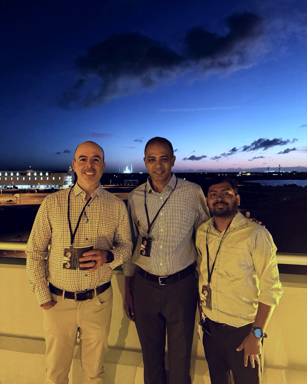 Three people wearing conference badges pose on a balcony at sunset, with a launch complex and lit buildings visible on the horizon.