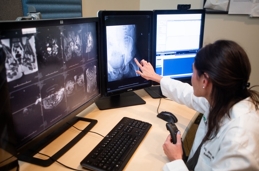 The back of a radiologist pointing to the computer screen with several monitors around her. She has scans up on each monitor and is holding a clicker.