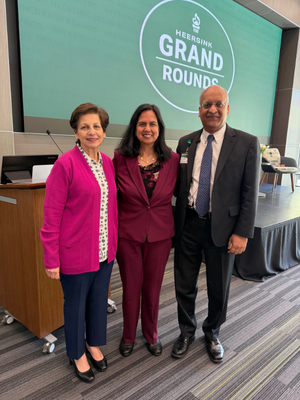Mona, Fouad, M.D., MPH, Sharmila Makhija, M.D., MBA, and Anupam Agarwal, M.D., stand in front of a large green graphic behind them displays "Heersink Grand Rounds" in white text with a white torch and a white circle.