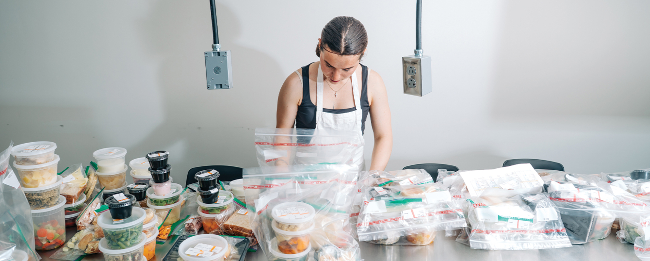 A registered dietitian is wearing apron and food service gloves while packaging food at counter of the UAB Teaching Kitchen.