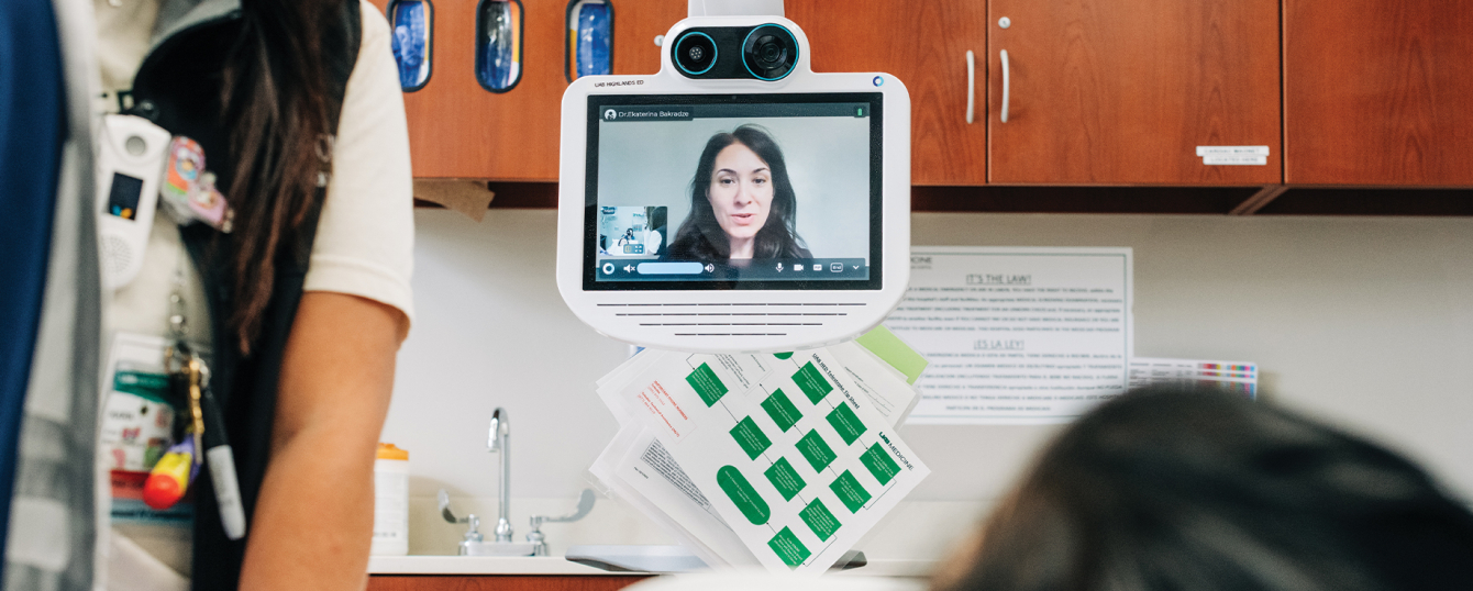 A female healthcare worker standing beside a hospital bed while speaking with a staged teleneurology (telestroke) female patient in an examination room while Dr. Ekaterina Bakradze, MD (Associate Professor, Neurology) is watching and speaking on a computer screen in the background.