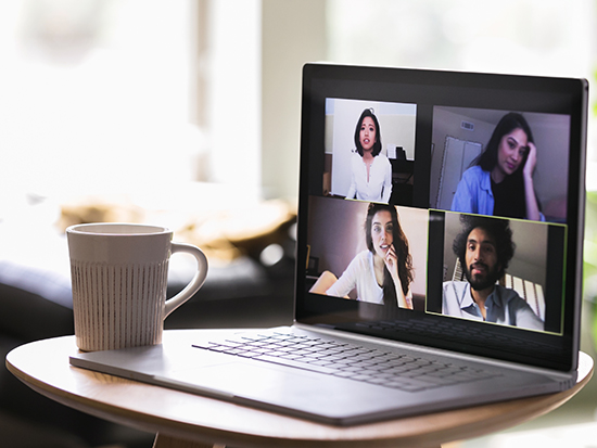 A group of young adults talk with one another virtually during the COVID-19 quarantined. Their images are seen on a laptop. The laptop is on a coffee table in a living room.