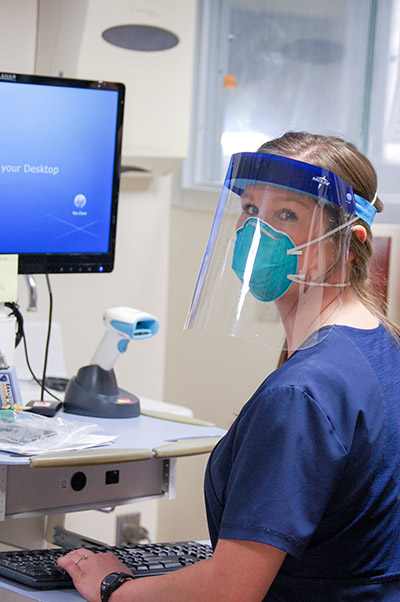 From side, an unidentified female medical professional is wearing a PPE (Personal Protective Equipment) face mask and shield while working at a computer in UAB Hospital during COVID-19 (Coronavirus Disease) pandemic, April 2020.