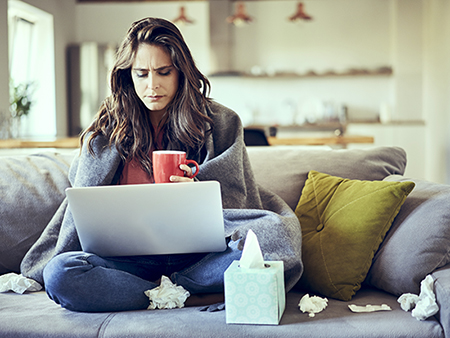 Sick woman sitting on sofa covered in blanket with cup of tea and laptop