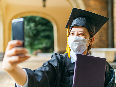 A student in graduation gown is holding her diploma and taking selfie with a smart phone.