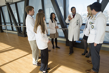 Dr. Selwyn Vickers, MD (Senior VP, Medicine; Dean, School of Medicine) is wearing white medical coat talking to several medical students, also wearing white medical coats, while standing on the 3rd Floor Breezeway of the North Pavilion, 2015.
