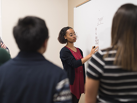 A young ethnic female professor stands at a large whiteboard and writes down Spanish verbs to conjugate. The shot is over the shoulders of several students.