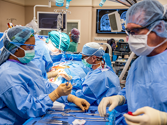 From side, Gary Roberts (Surgical Tech, Perioperative Services), two female health care workers, and Dr. James Markert, MD (Professor and Chair, Neurosurgery) are wearing PPE (Personal Protective Equipment) gowns, head covers, face masks, and gloves while performing surgery on a brain tumor in an operating room in UAB Hospital, 2020. 