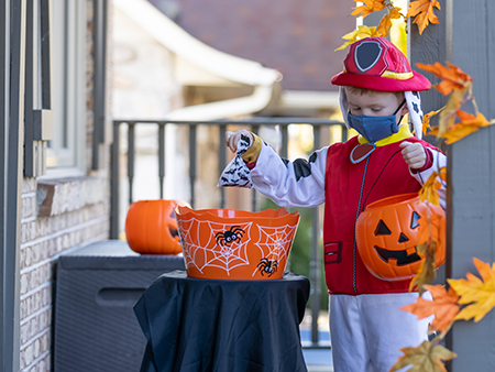 Young Boy wearing protective mask taking candies in a self service candy basket on Halloween at front door of a house during COVID-19 pandemic. People are putting a candy basket outside to keep social distancing and let children celebrate Halloween and trick or treat.