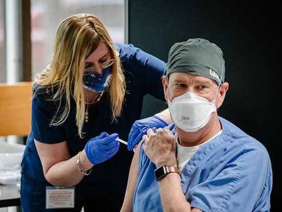 Dr. David Pigott, MD (Professor, Emergency Medicine) is wearing blue scrubs and a PPE (Personal Protective Equipment) head cover, and face mask while receiving the COVID-19 (Coronavirus Disease) vaccination from a female healthcare worker wearing scrubs and a PPE face mask and gloves, December 2020.
