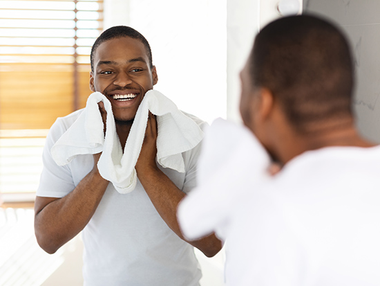 Aftershave Care Concept. Handsome Black Guy Wiping Face With Towel In Bathroom, Young African American Man Looking At Mirror And Smiling While Making Morning Hygiene, Selective Focus On Reflection