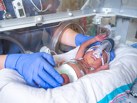 Close-up, nurse's hands wearing blue safety gloves are tending to a premature baby in a neonatal incubator in the Regional Neonatal Intensive Care Unit (NICU), 2019.