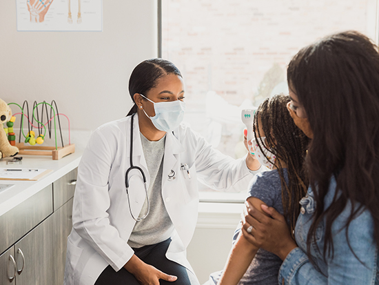 During COVID-19, everyone wears protective masks during the doctor's examination.  The doctor takes the girl's temperature with a touch free thermometer.