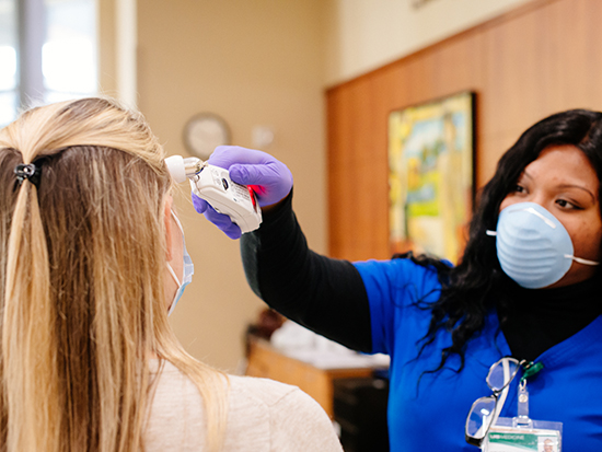 A black health care worker is wearing a PPE (Personal Protective Equipment) face mask and gloves while taking the temperature of a female patient visiting a medical office in the Women and Infants Center during COVID-19 (Coronavirus Disease), May 2020.