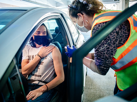 From side, an unidentified female health care worker is wearing a safety vest PPE (Personal Protective Equipment) gloves and face mask is administering the influenza vaccination to a male patient wearing a face mask and sitting in a parked automobile at the drive-thru flu shot event in the 12th Street Parking Deck at the Community Health Services Building during the COVID-19 (Coronavirus Disease) pandemic, November 2020.  