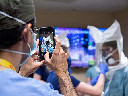 Back, Dr. Nitin Arora, MD (Assistant Professor, Pediatrics - Neonatology) is holding a cell phone/mobile phone of while taking a digital photograph of Dr. Albert Pierce II, (Professor, Anesthesiology) while he is being fitted with a respirator that was adapted to include a medical grade filter at a demonstration testing the fit of PPE (Personal Protective Equipment) mask at the UAB Hospital COVID-19 (Coronoavirus Disease) Command Center in the North Pavilionon on April 14, 2020.