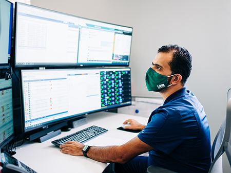 From side, a male health care worker is wearing a face mask due to the COVID-19 (Coronavirus Disease) pandemic while looking at computer monitors while working in the UAB Tele-ICU Operations Center, a partnership between UAB and Advanced ICU Care to provide physician services and consulatation services to ICUs across Alabama, September 2021.