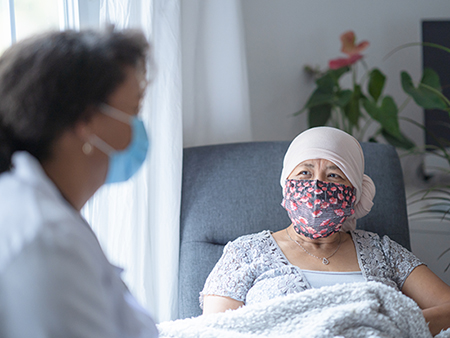 In a hospital room, an asian female cancer patient is chatting with her doctor. Both of them are wearing a mask during the coronavirus pandemic to help prevent the transfer of germs. The doctor is a female and of African American ethnicity.