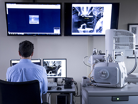 Back, man is sitting in chair in front of computer monitors beside microscope in the School of Engineering Scanning Electron Microscopy Laboratory, 2018.